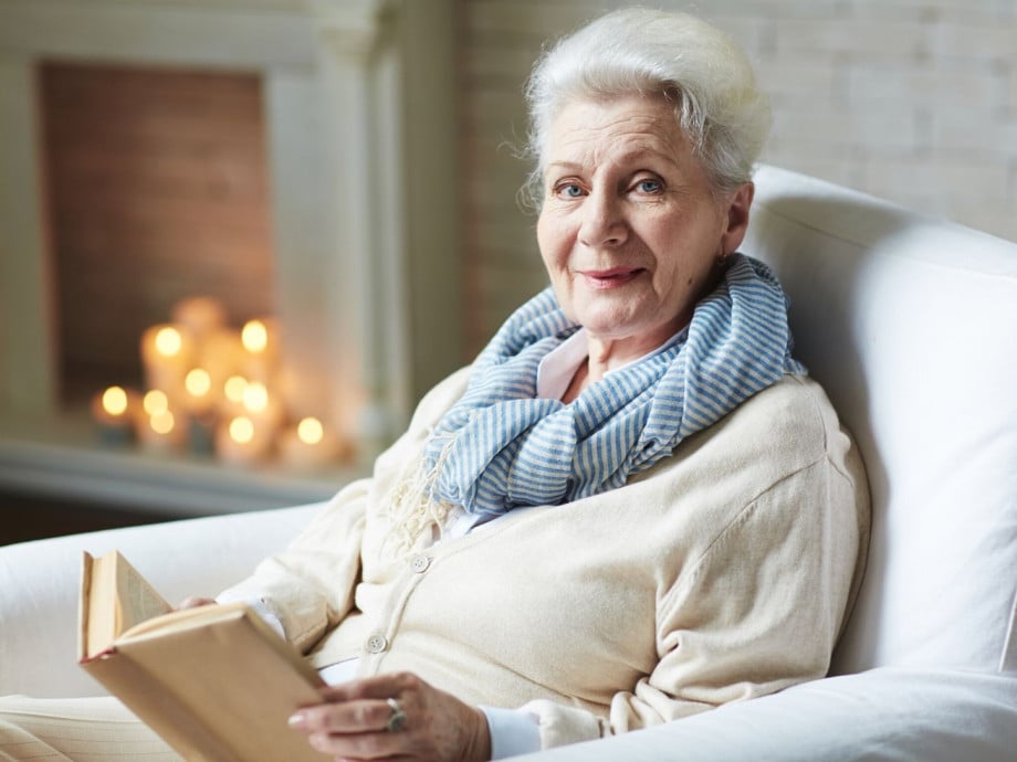 White-haired woman sitting in front of the fireplace with a book.