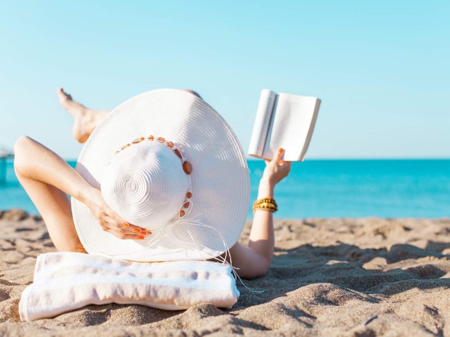 Woman with sun hat lying on the beach, reading a book.