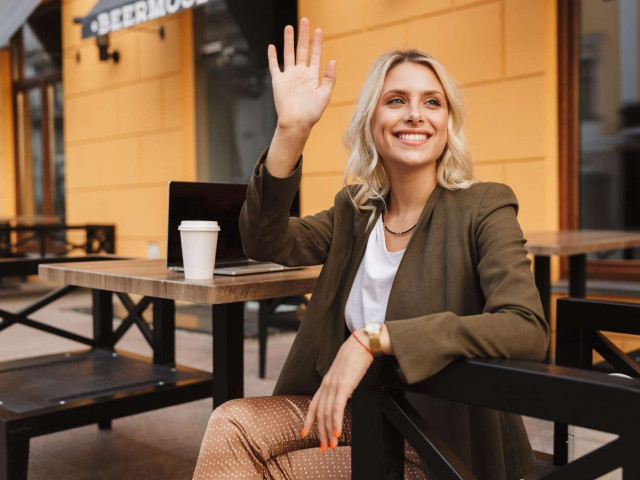 Woman sitting in a café waving hello to someone outside the frame